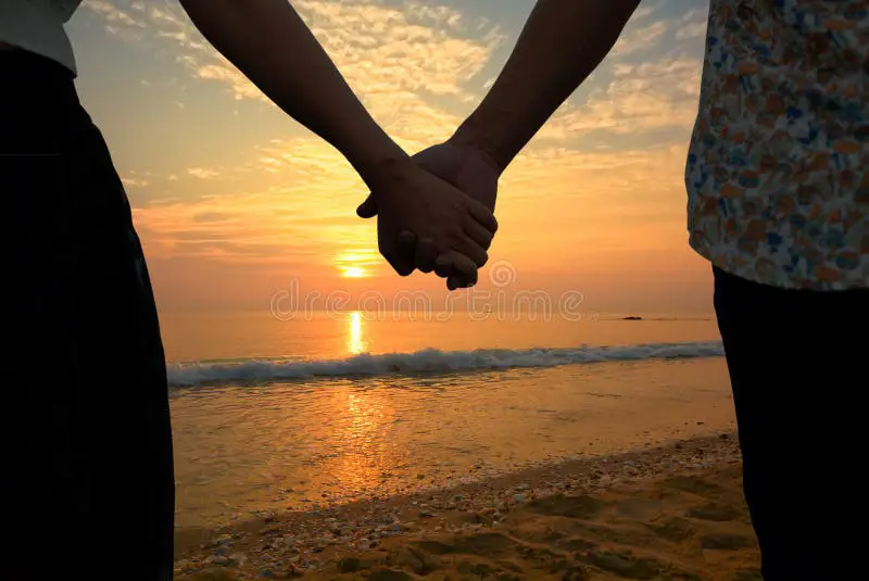 Pasangan memegang tangan di pantai saat matahari terbenam